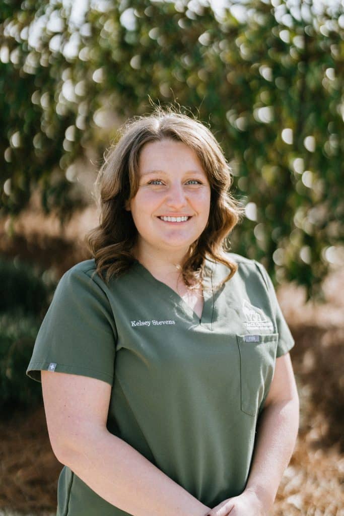 Headshot of Kelsey Stevens, Practice Manager. Kelsey is standing in front of a blurred out Magnolia tree. Kelsey is standing at an angle and the picture is from her waist up.